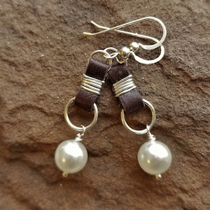 May include: A pair of dangle earrings featuring silver-tone hooks and hoops. Each earring has a dark brown leather accent wrapped with silver wire, and a large, pearlescent white bead. The earrings are displayed on a brown, textured surface.