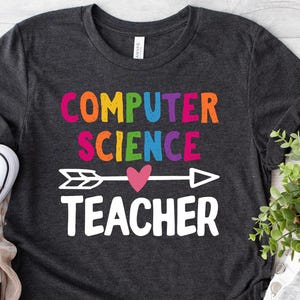Puede incluir: Camiseta gris oscuro con las palabras "COMPUTER SCIENCE TEACHER" en texto colorido y una flecha blanca con un corazón rosa. Zapatillas de lona rosas con cordones blancos junto a la camiseta.