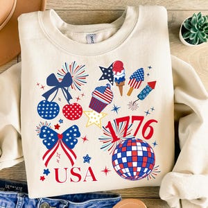 May include: Cream-colored sweatshirt with a patriotic design featuring red, white, and blue graphics. The design includes fireworks, stars, bows, and the text "USA" and "1776". A hat and jeans are visible in the background.