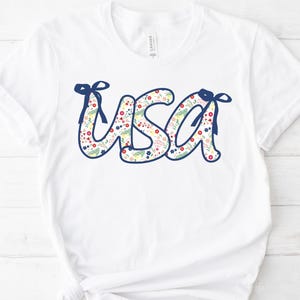 Peut inclure: T-shirt blanc avec le mot "USA" dans un motif floral. Les lettres sont soulignées en bleu marine et ornées de nœuds bleu marine. Le motif floral comprend des fleurs rouges, roses et jaunes avec des feuilles vertes.