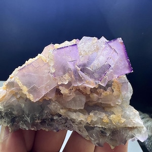 May include: A natural fluorite mineral specimen with a mix of colors. The top features vibrant purple, square-shaped crystals. The base has a mix of light purple, clear, and tan crystals. The overall structure is rough and irregular, showcasing natural formations.
