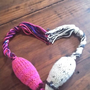 May include: Two knitted cat toys shaped like mice, connected in a heart shape. One mouse is bright pink, the other is off-white with black flecks. The tails are twisted yarn in shades of pink, purple, and white.