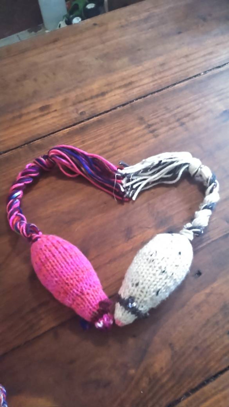 May include: Two knitted cat toys shaped like mice, connected in a heart shape. One mouse is bright pink, the other is off-white with black flecks. The tails are twisted yarn in shades of pink, purple, and white.