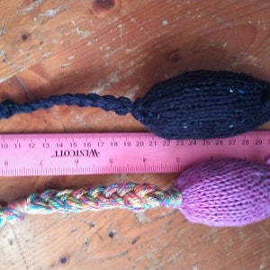 May include: Two knitted cat toys, one black and one purple, with braided tails. The black toy is approximately 4 inches long, and the purple toy is about 5 inches long. A pink ruler is placed between the toys.