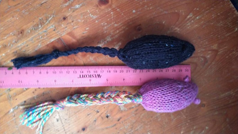May include: Two knitted cat toys, one black and one purple, with braided tails. The black toy is approximately 4 inches long, and the purple toy is about 5 inches long. A pink ruler is placed between the toys.