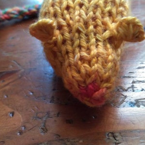 May include: Close-up of a knitted toy mouse in shades of yellow and orange. The mouse has small, rounded ears and a pink nose. A multicolored yarn tail extends from the back. The toy is handmade and sits on a wooden surface.