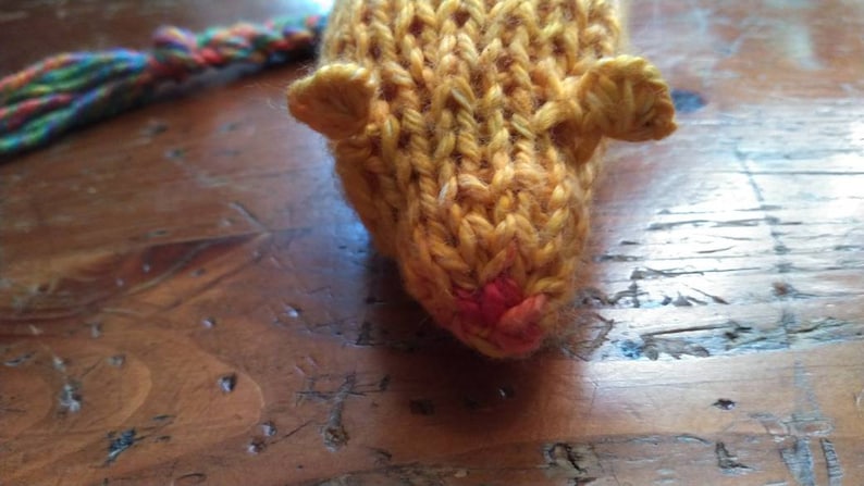 May include: Close-up of a knitted toy mouse in shades of yellow and orange. The mouse has small, rounded ears and a pink nose. A multicolored yarn tail extends from the back. The toy is handmade and sits on a wooden surface.