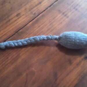 May include: A gray knitted cat toy shaped like a mouse, with a braided tail and a pink nose. The toy is lying on a wooden surface. The tail is made of the same gray yarn as the body.