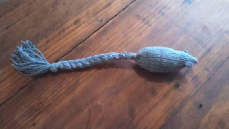 May include: A gray knitted cat toy shaped like a mouse, with a braided tail and a pink nose. The toy is lying on a wooden surface. The tail is made of the same gray yarn as the body.