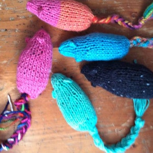 May include: A collection of five handmade knitted cat toys shaped like mice. The toys are in various colors, including pink, blue, teal, black, and orange. Each mouse has a braided yarn tail in coordinating colors. These toys are designed for pet play.