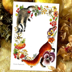 May include: A rectangular white card featuring a watercolour illustration of a badger and a fox surrounded by a floral border of red berries, yellow flowers, white blossoms, purple pansies, and a red-spotted mushroom. The text Giulia Pagliarulo is printed in the bottom left corner.