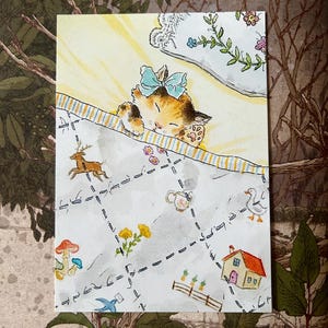 May include: A rectangular art print featuring a watercolour illustration of a calico kitten with a light blue bow sleeping under a striped blanket. The blanket is decorated with small drawings of a deer, a house, a duck, mushrooms, and flowers.