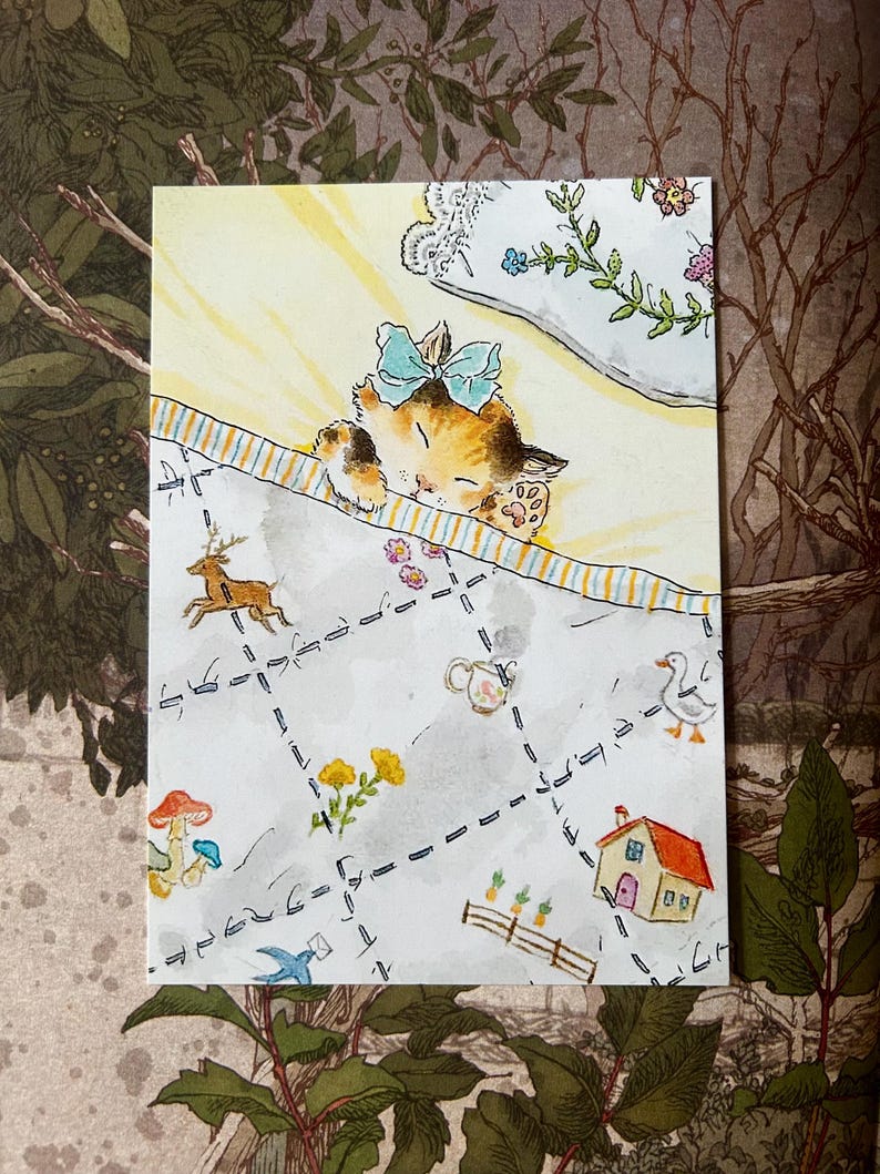 May include: A rectangular art print featuring a watercolour illustration of a calico kitten with a light blue bow sleeping under a striped blanket. The blanket is decorated with small drawings of a deer, a house, a duck, mushrooms, and flowers.