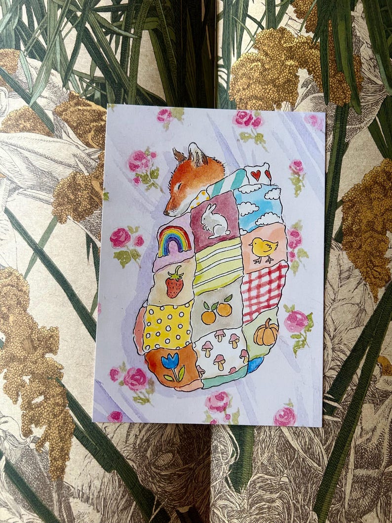 May include: A rectangular art print featuring a watercolour illustration of a fox sleeping under a patchwork quilt. The quilt is composed of various squares displaying motifs including a rainbow, a rabbit, a chick, a strawberry, mushrooms, and a pumpkin.