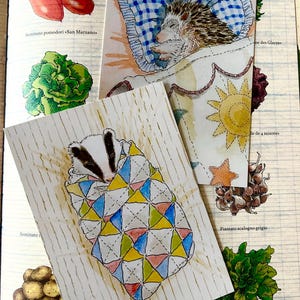 May include: Two rectangular art prints featuring watercolour illustrations of animals sleeping. One print shows a hedgehog in a blue checkered bed, the other shows a badger wrapped in a colourful geometric quilt, both resting on a page with vegetable drawings.