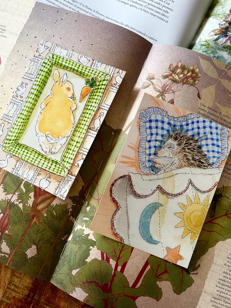 May include: Two rectangular art prints featuring watercolour illustrations. One print shows a yellow rabbit sleeping on a green checkered mat with a carrot. The other print depicts a hedgehog sleeping on a blue checkered pillow under a blanket with a moon, sun, and star.