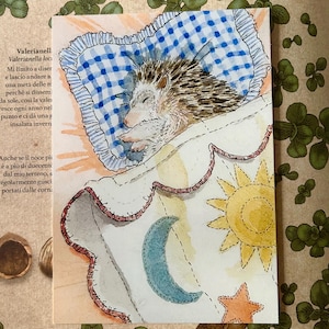 May include: A watercolour illustration of a hedgehog sleeping on a blue and white checkered pillow. The hedgehog is tucked under a white blanket decorated with a yellow sun, a blue crescent moon, and an orange star, featuring a red stitched edge.