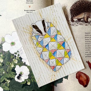May include: A rectangular card featuring a watercolour illustration of a badger wrapped in a quilt with a geometric pattern of blue, yellow, and pink triangles. The card has a white background with thin vertical gold lines and is placed on a book page.