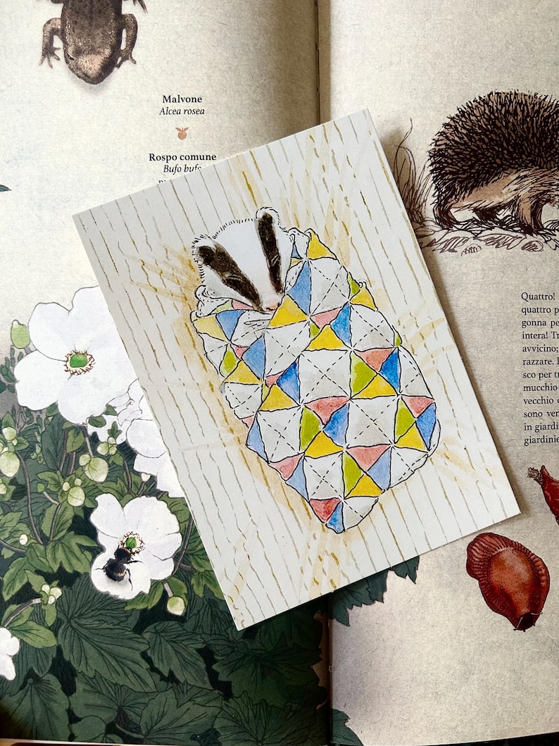 May include: A rectangular card featuring a watercolour illustration of a badger wrapped in a quilt with a geometric pattern of blue, yellow, and pink triangles. The card has a white background with thin vertical gold lines and is placed on a book page.