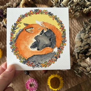 May include: Watercolour painting of a fox and a wolf embracing, encircled by a wreath of berries and leaves. The fox is orange, the wolf grey, set against a yellow background. The artwork is square.