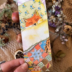 May include: A watercolour bookmark depicting a sleeping fox with orange fur, under a floral pillow. The bottom section shows a colourful patchwork quilt design, creating a cosy and whimsical aesthetic.