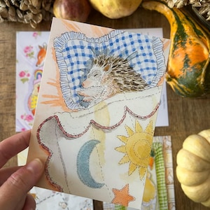May include: A watercolour illustration on a rectangular card depicting a hedgehog sleeping on a blue and white chequered pillow. The hedgehog is covered by a white blanket featuring a yellow sun, a blue crescent moon, and an orange star.