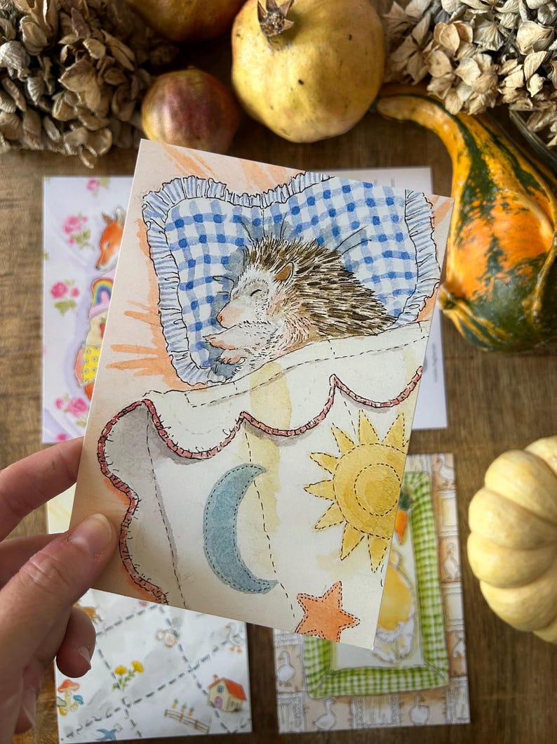 May include: A watercolour illustration on a rectangular card depicting a hedgehog sleeping on a blue and white chequered pillow. The hedgehog is covered by a white blanket featuring a yellow sun, a blue crescent moon, and an orange star.