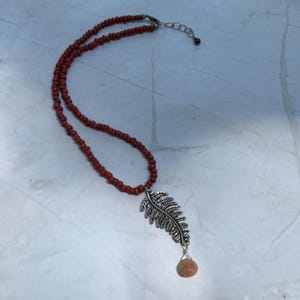 May include: A red beaded necklace featuring a silver-coloured fern pendant and a small, peach-coloured stone. The necklace is displayed on a light-coloured surface.