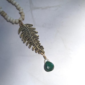 May include: A gold-coloured fern pendant necklace with a teardrop-shaped green gemstone. The necklace has a beaded strand of white and tan beads. The pendant is detailed with intricate leaf veins.