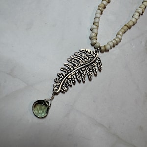 May include: A silver fern leaf pendant necklace with a green gemstone drop. The pendant is attached to a beaded necklace with off-white beads. The necklace is displayed on a light-coloured surface.