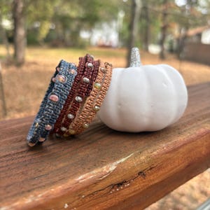 Può includere: Tre braccialetti in macramè blu, rosso scuro e marrone chiaro, ognuno con un motivo a corda intrecciata con perline metalliche e di vetro incastonate. I braccialetti sono impilati accanto a una piccola zucca decorativa bianca.