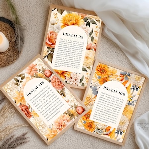 May include: Three framed floral art prints featuring Bible verses Psalm 23, Psalm 118, and Psalm 100. The prints have a beige background with watercolor flowers in shades of yellow, orange, and white. Each print is in a light brown wooden frame.
