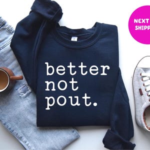 May include: A navy blue sweatshirt with the words "better not pout" in white block letters. The sweatshirt is displayed with a pair of light blue jeans, a brown mug, and gray sneakers.