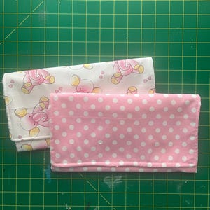 以下が含まれることがあります： ピンクと白のドット柄の生地と、白地にピンクの象と黄色のアクセントとピンクのハートが描かれた生地の2枚の正方形の生地です。