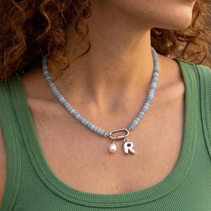 May include: A beaded necklace featuring light blue spherical beads, a silver-toned oval carabiner clasp, a single white pearl drop charm, and a silver-toned letter R charm. The necklace is worn around the neck against a green ribbed tank top.