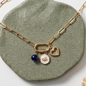 May include: Gold-toned chain necklace with a large clasp and three charms. Charms include a blue bead, a white pendant with a fish design, and a heart. The necklace is displayed on a green, textured surface with a starfish and seashell.