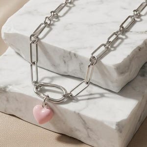 May include: A silver-toned chain necklace featuring elongated rectangular links and a single, smooth, light pink heart-shaped pendant attached to a larger, rounded oval link at the center of the chain, creating a simple and balanced design.