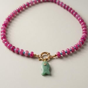 May include: A beaded necklace featuring small, round, faceted magenta-pink beads interspersed with tiny turquoise spacers. The necklace includes a gold-toned circular clasp with a light green, fish-shaped stone pendant featuring a small embedded gem.