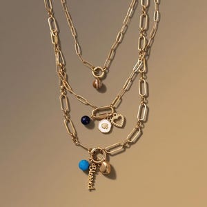 May include: Three gold-colored chain necklaces with various charms. The charms include a heart, a blue bead, a gold-colored bell, a white pendant with a gold design, and a charm that says "Blessed". The necklaces are layered.
