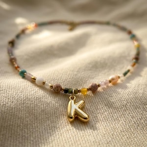 May include: A beaded necklace featuring a variety of small, multi-colored gemstone beads in shades of pink, blue, yellow, and white, accented with gold-toned spacers and a central, polished gold-toned letter K pendant charm.