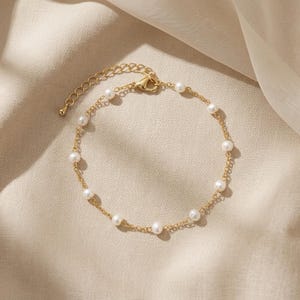 May include: A delicate gold-toned bracelet featuring a series of small, round, white pearls. The bracelet has a fine chain design with a lobster clasp closure and an adjustable chain for a comfortable fit. The jewelry is displayed on a neutral-toned fabric.