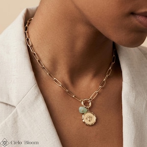 May include: Gold-toned chain necklace with a large link design. The necklace features a round pendant with a bee design and a small, light green stone. The necklace is worn on a person wearing a light-colored blazer.