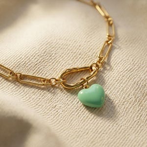May include: A gold-toned chain necklace featuring elongated rectangular links and a central, irregularly shaped, textured gold-toned clasp. A smooth, solid mint green heart-shaped pendant hangs from the clasp, creating a distinct contrast.