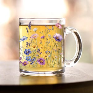 May include: A clear glass mug with a handle, filled with light yellow tea. The mug has a floral design featuring purple, pink, yellow, and blue flowers.