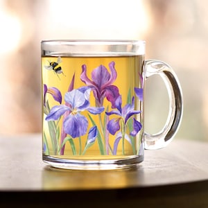 May include: A clear glass mug with a handle features a colorful floral design of purple and blue irises with green leaves. A small yellow and black bee is flying above the flowers.