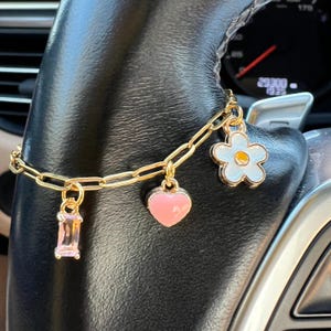 May include: Gold-toned charm bracelet featuring a rectangular pink gemstone, a pink heart, and a white and blue flower charm. The bracelet is displayed on a black surface.