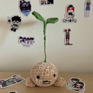 Puede incluir: Un brote de peluche de ganchillo con una cara sonriente, cuerpo marrón claro y hojas verdes. El brote está rodeado de pegatinas con la palabra "OMORI" e ilustraciones de personajes.