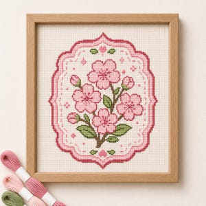 以下が含まれることがあります： クリーム色の布地にピンクの桜の花と緑の葉を中央に配置し、ピンクのスカラップ模様の縁取りで囲んだクロスステッチ刺繍の額装品。刺繍糸の束が数本添えられています。