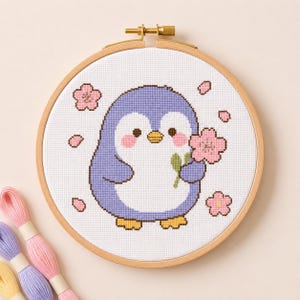 Op de afbeelding: Een ronde houten borduurring met een kruissteekontwerp van een lichtpaarse pinguïn met een witte buik, roze wangen en gele voeten, die een roze kersenbloesem vasthoudt, omringd door verspreide roze bloemblaadjes op witte stof.