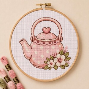 May include: A cross-stitch embroidery project in a round wooden hoop featuring a light pink teapot with white polka dots, a heart-shaped lid knob, and a cluster of white flowers with green leaves at the base, accompanied by three pink embroidery floss skeins.